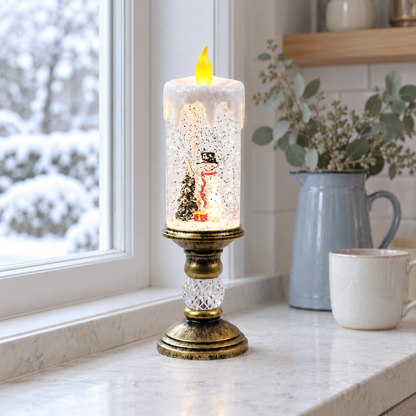 Lit snow globe candle with a snowman in a top hat and red scarf beside a tree and a red gift, on a marble kitchen windowsill with snow outside, beside a blue jug and a mug