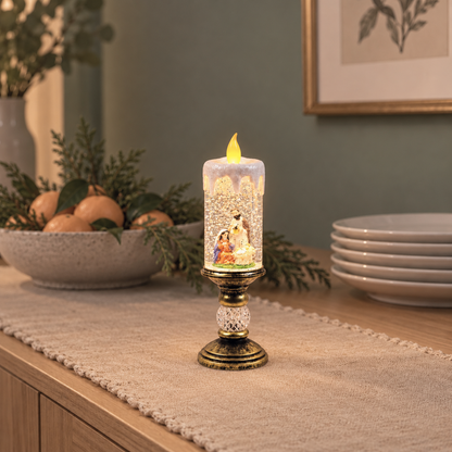 Lit snow globe candle with the nativity scene of Mary, Joseph and the baby, on a linen runner on an oak dining sideboard beside a bowl of clementines and a stack of plates