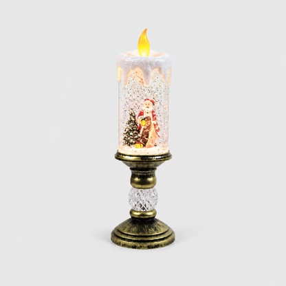 Christmas snow globe candle lit on a light gray background, Santa in a long tan robe and red coat holding a lantern beside a snowy tree inside the glittering column, on an antique gold-look pedestal