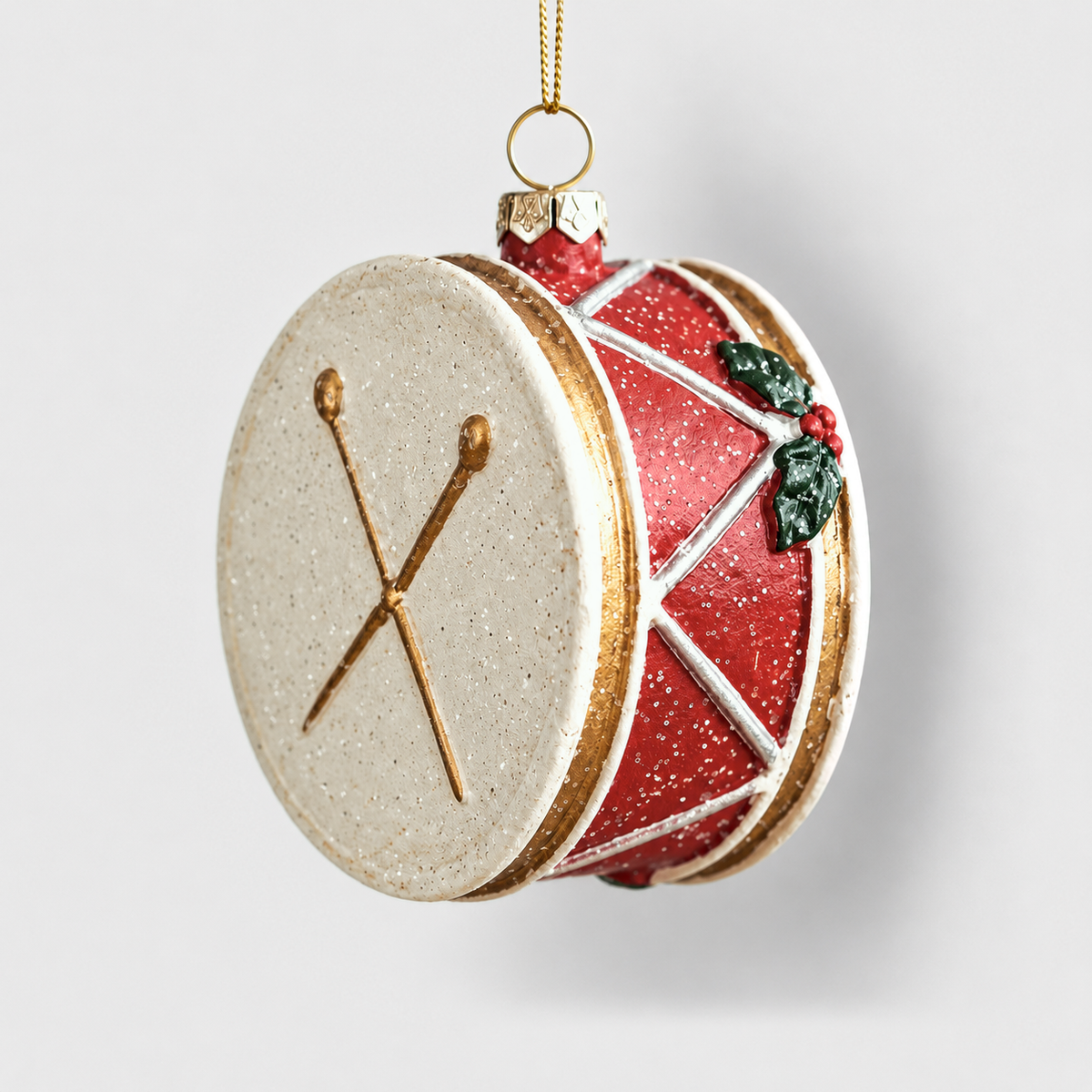 Red Christmas drum ornament with a cream head, crossed gold drumsticks and white zigzag cords, on a light gray background