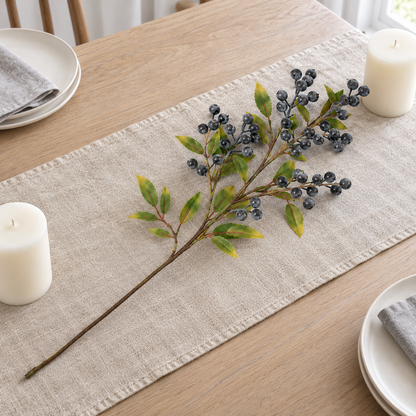 Blue berry branch lying along a linen table runner between white pillar candles