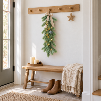 Short green pine swag with a cream bow, small baubles and fairy lights hanging from a wooden peg in a white hallway