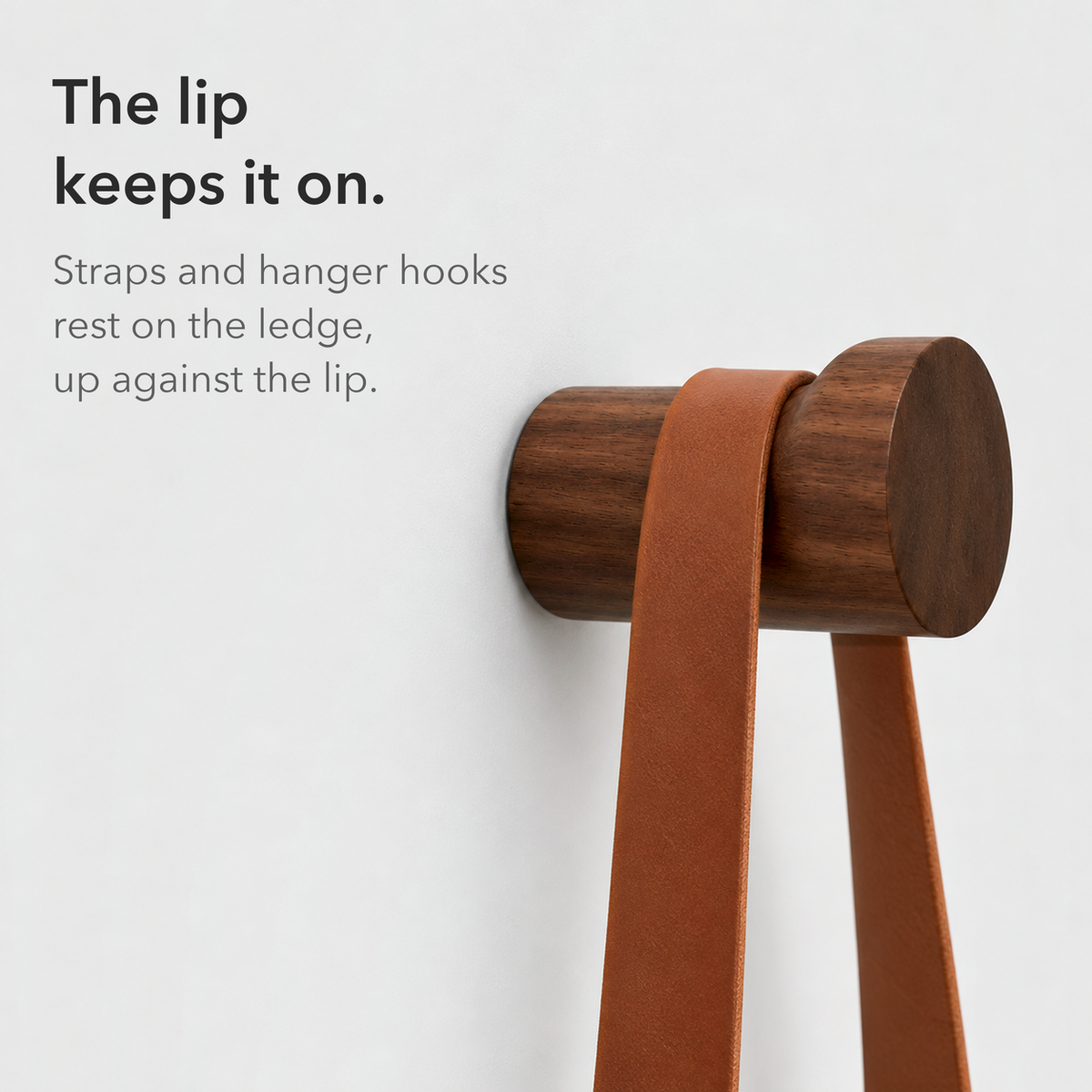 Close side view of a walnut wall hook with a leather strap resting against its raised lip, text reading the lip keeps it on