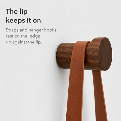 Close side view of a walnut wall hook with a leather strap resting against its raised lip, text reading the lip keeps it on