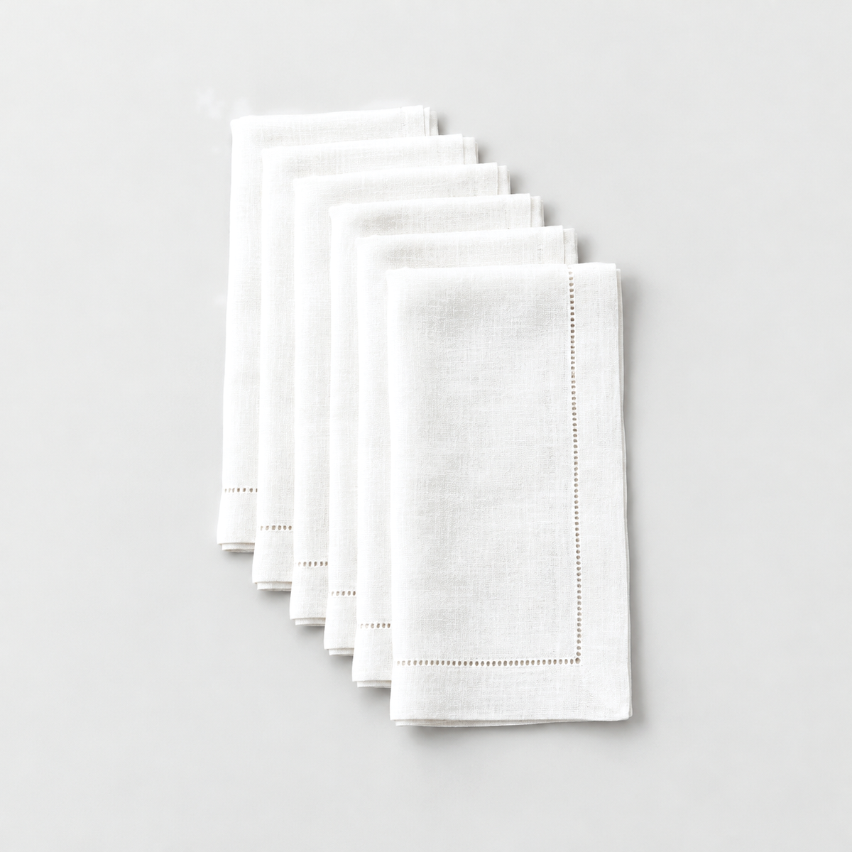 Six folded white hemstitch dinner napkins laid in a staggered row on a light grey background