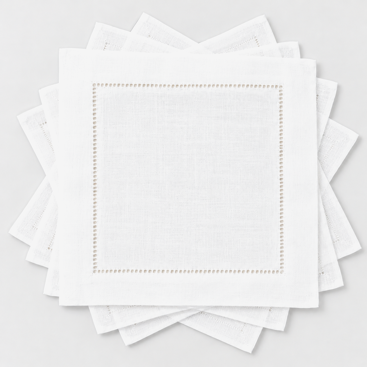 Six small white hemstitch cocktail napkins fanned out in a loose rotation on a light grey background