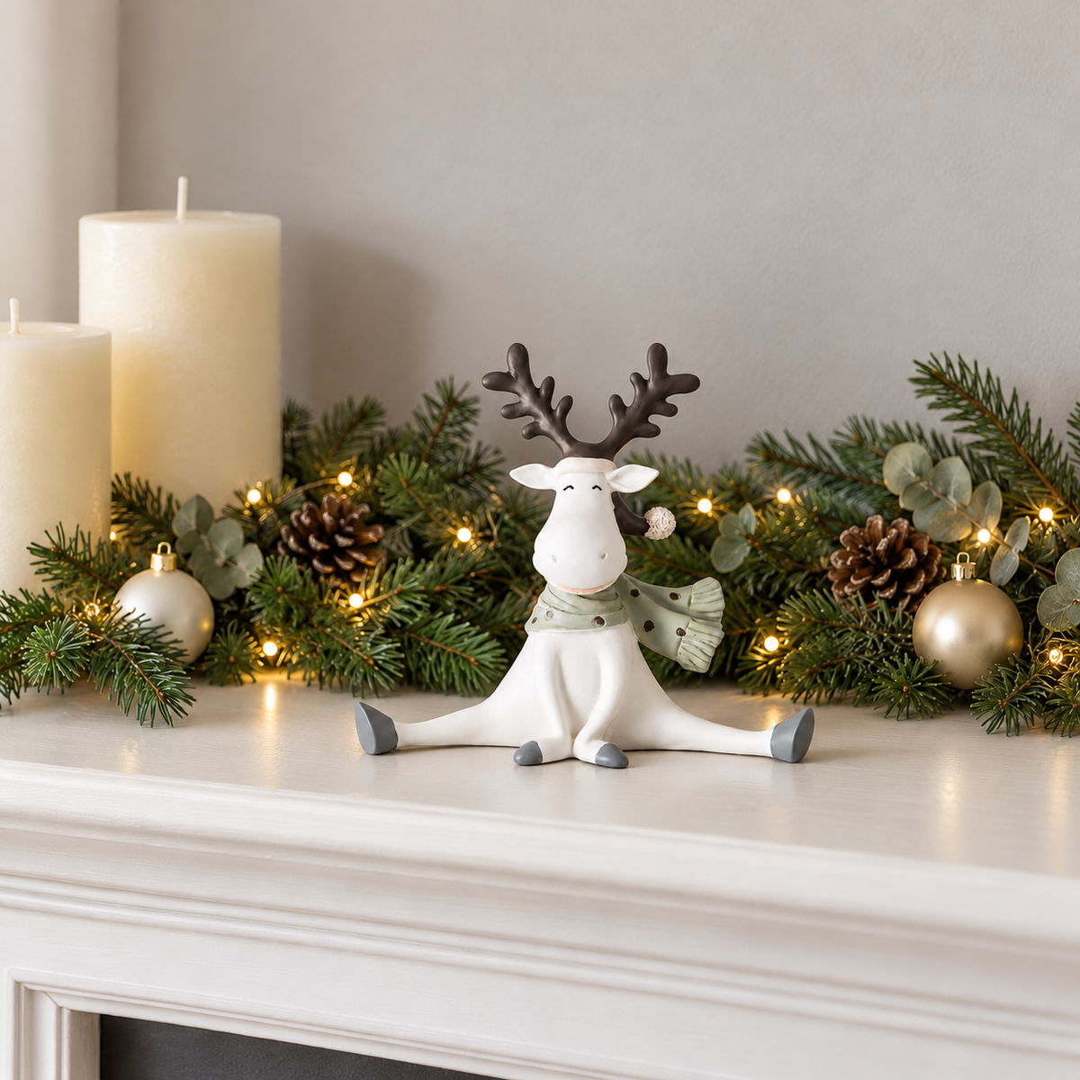 White resin reindeer figurine sitting in the splits on a white fireplace mantel in front of a fir garland with fairy lights