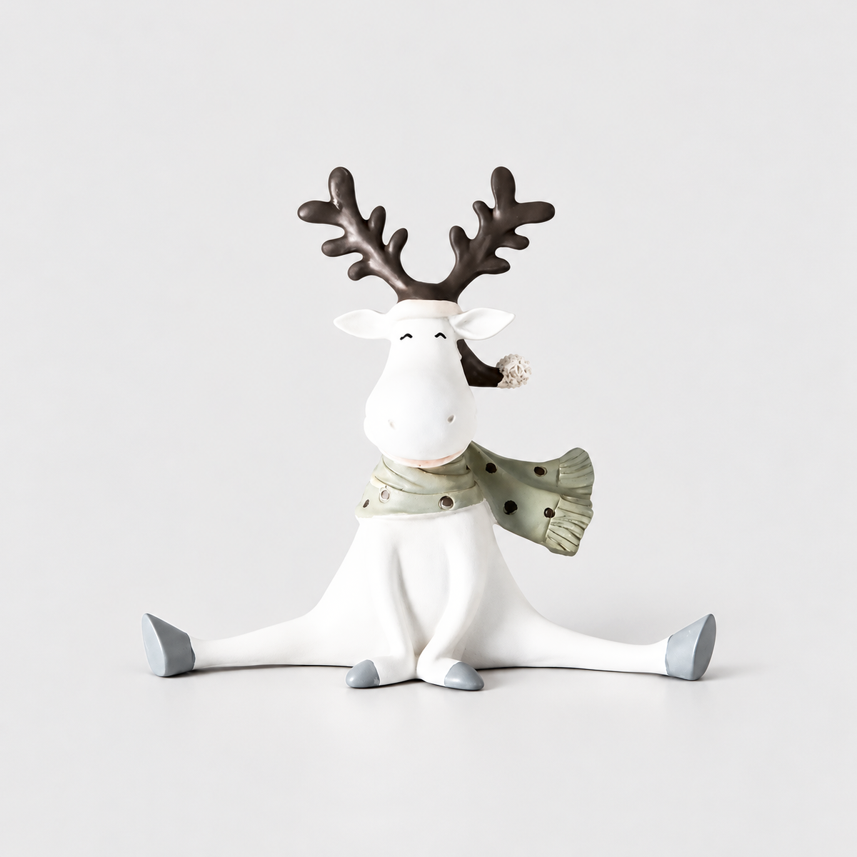 White reindeer figurine with brown antlers, a pompom hat and a green-gray dotted scarf, sitting with its legs spread on a light gray background