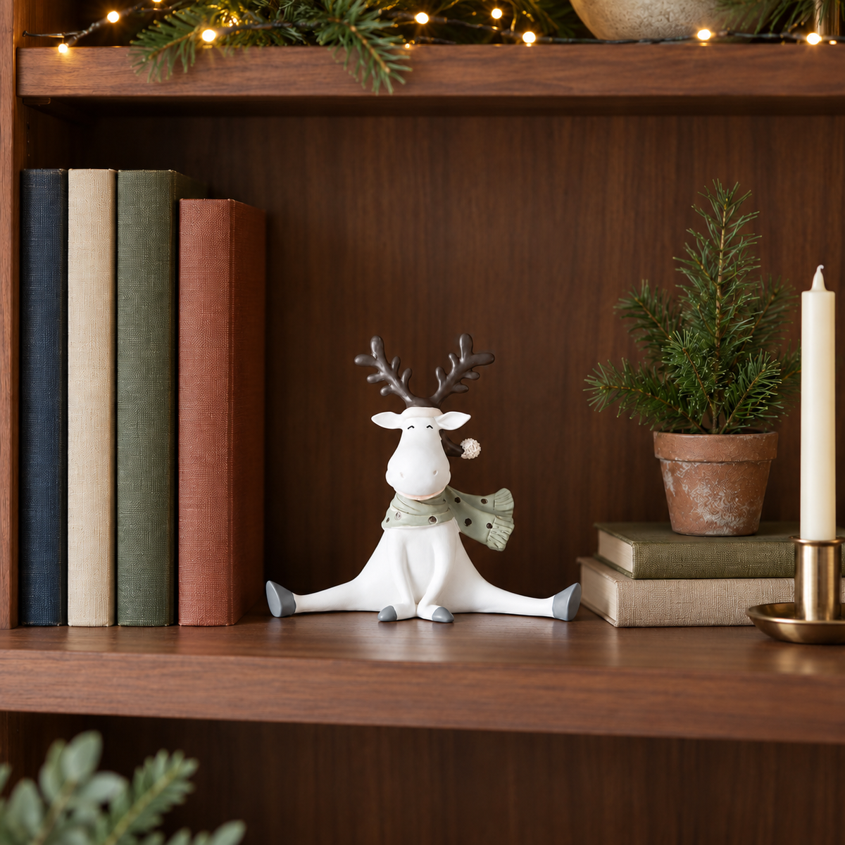 Reindeer figurine with a scarf sitting on a walnut bookshelf beside books and a small potted fir with fairy lights