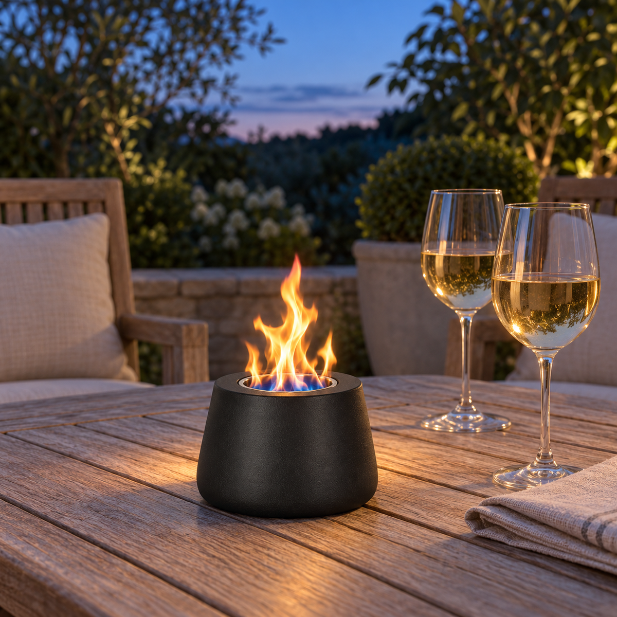 Black tabletop fire pit burning on a teak patio table at dusk beside two glasses of white wine