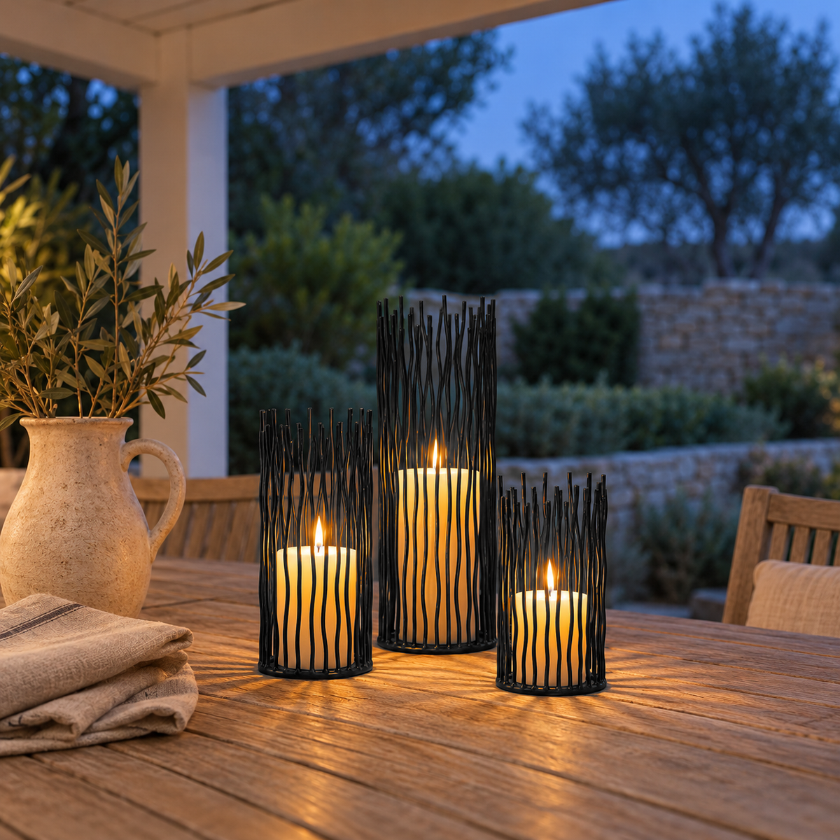 Three black iron candle holders with lit pillar candles on a teak table on a covered patio at dusk