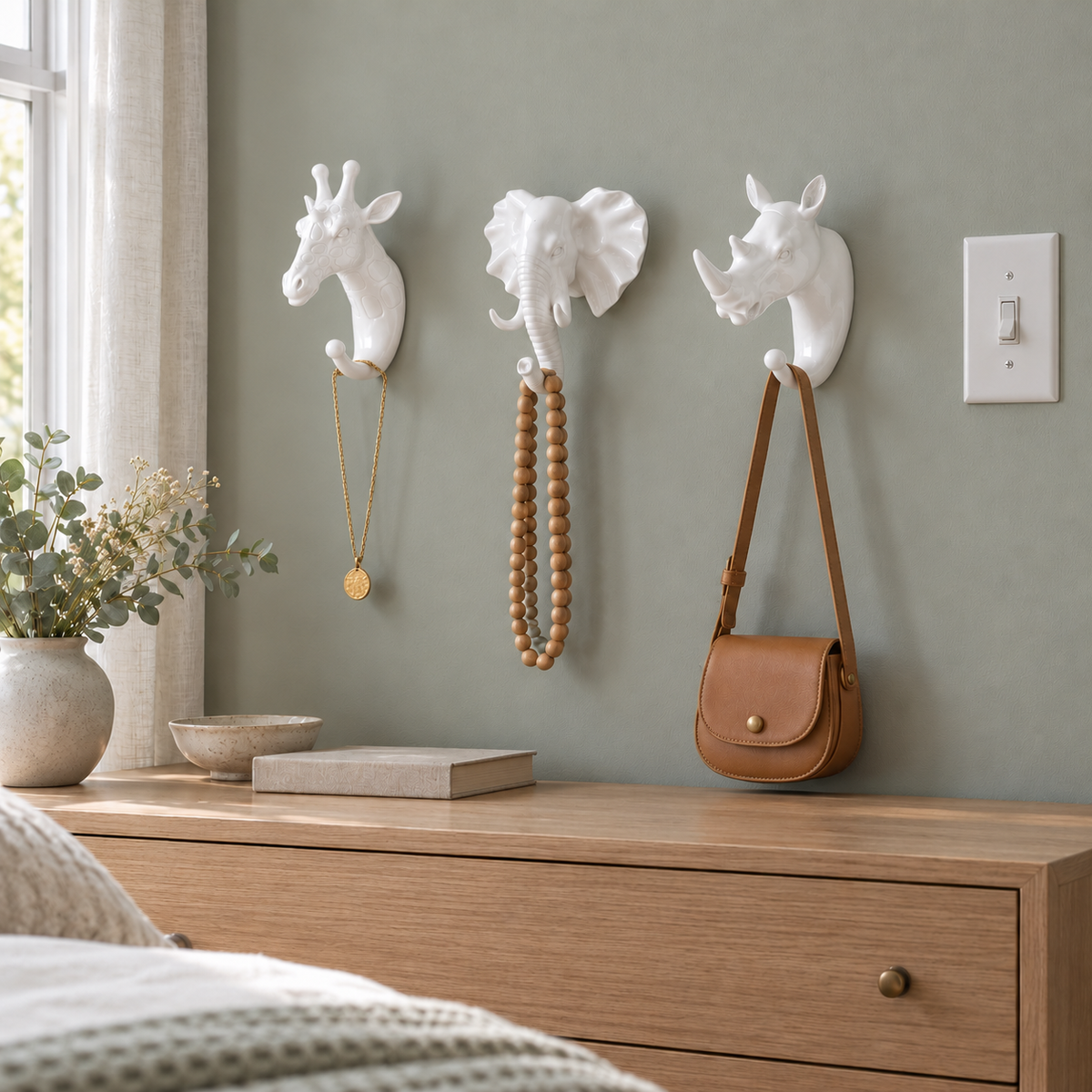 White giraffe, elephant and rhino head wall hooks on a sage gray bedroom wall holding necklaces and a small leather bag