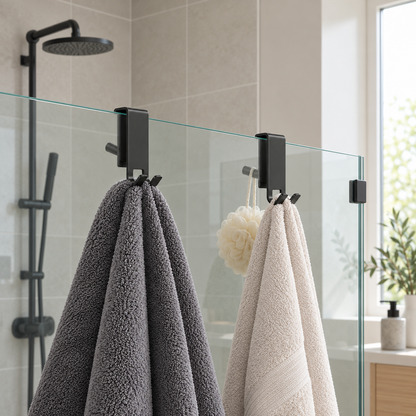 Two black shower door hooks on a frameless glass screen holding gray and stone towels, a loofah hanging from a peg inside
