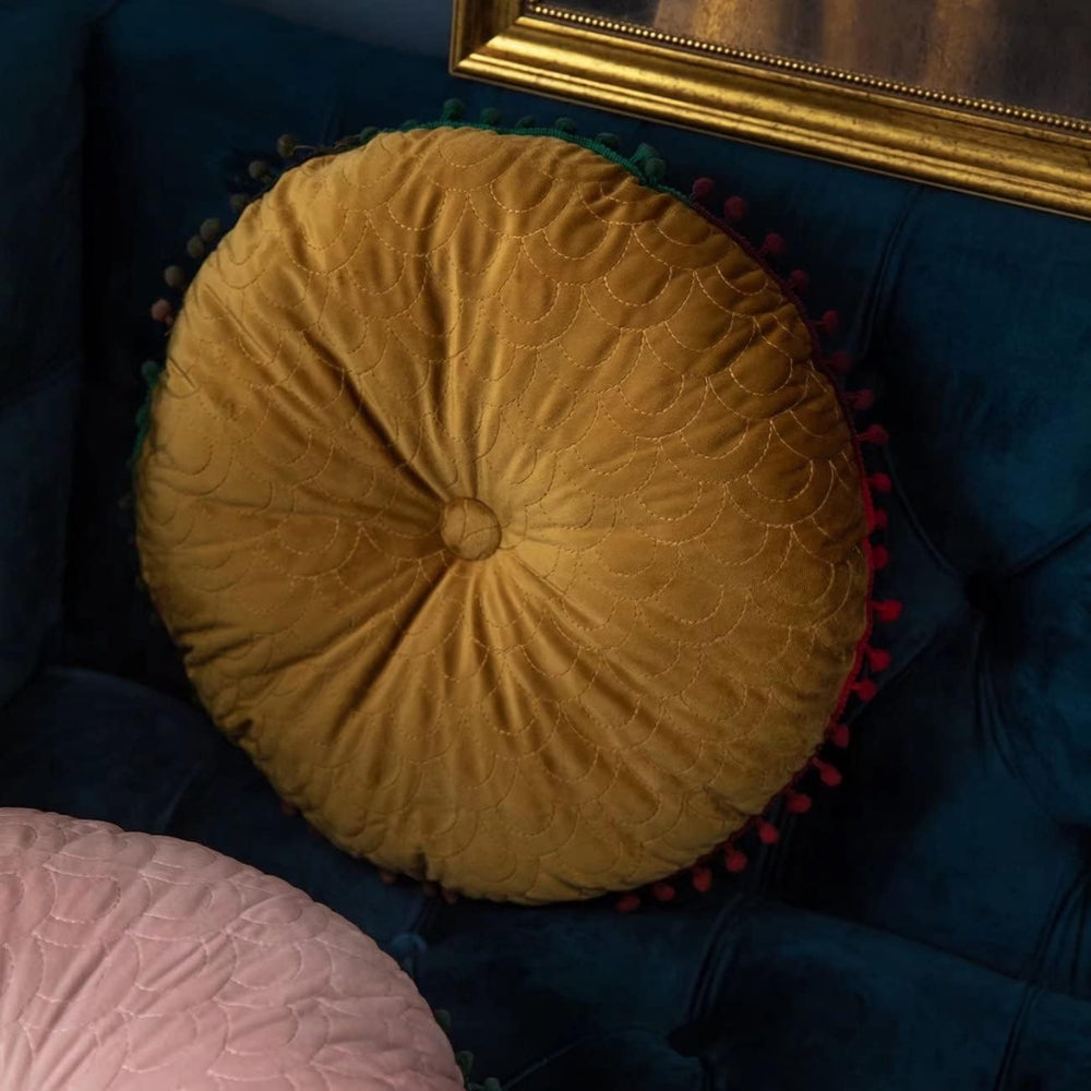 Mustard round velvet cushion leaning against a navy sofa, showing the button and scallop stitching