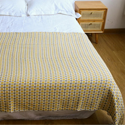 Mustard geometric knit throw spread across the end of a bed beside a rattan nightstand