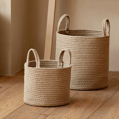 Small and medium tan cotton rope baskets with upright rope handles on a wood floor beside a pale wall