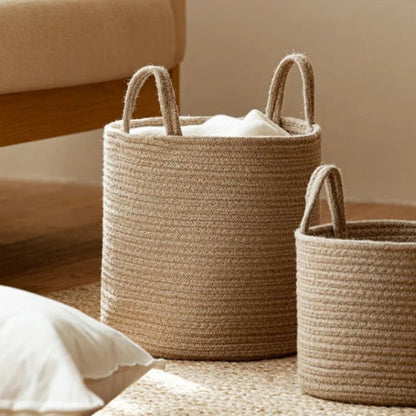 Tan cotton rope basket holding a white cloth on a cream rug, with a smaller basket and a pillow beside it