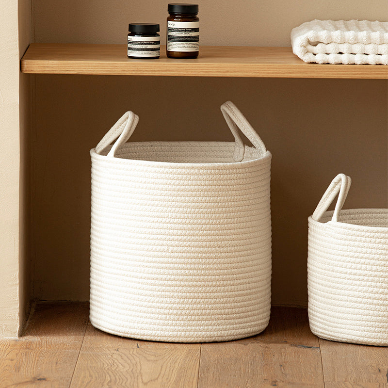 White cotton rope basket under a wooden shelf holding folded towels and two dark jars