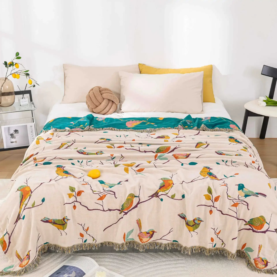 Cream cotton coverlet with songbirds on branches and a fringed edge spread over a bed in a bright white bedroom