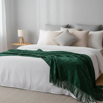 Deep green diamond knit throw blanket spread across a white bed with its tassel fringe hanging over the side
