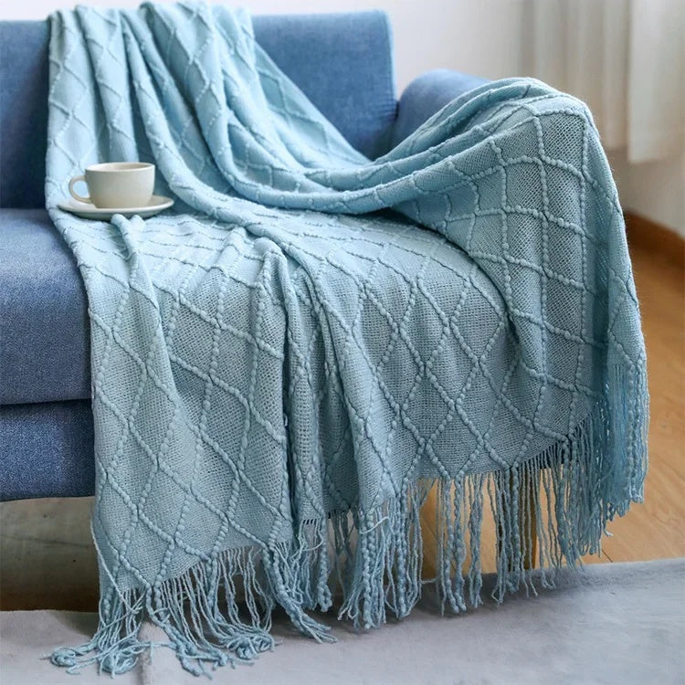 Pale blue knit throw blanket with raised diamond texture draped over a sofa with tassels on the floor