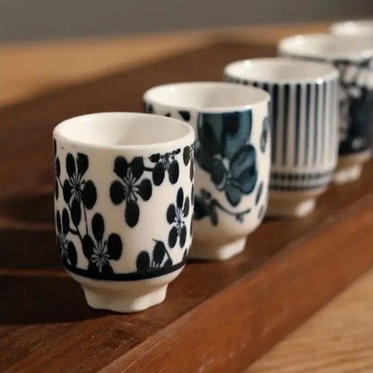 A close row of sake cups on a dark wooden board: blossoms, a brushed leaf and fine stripes in deep blue.