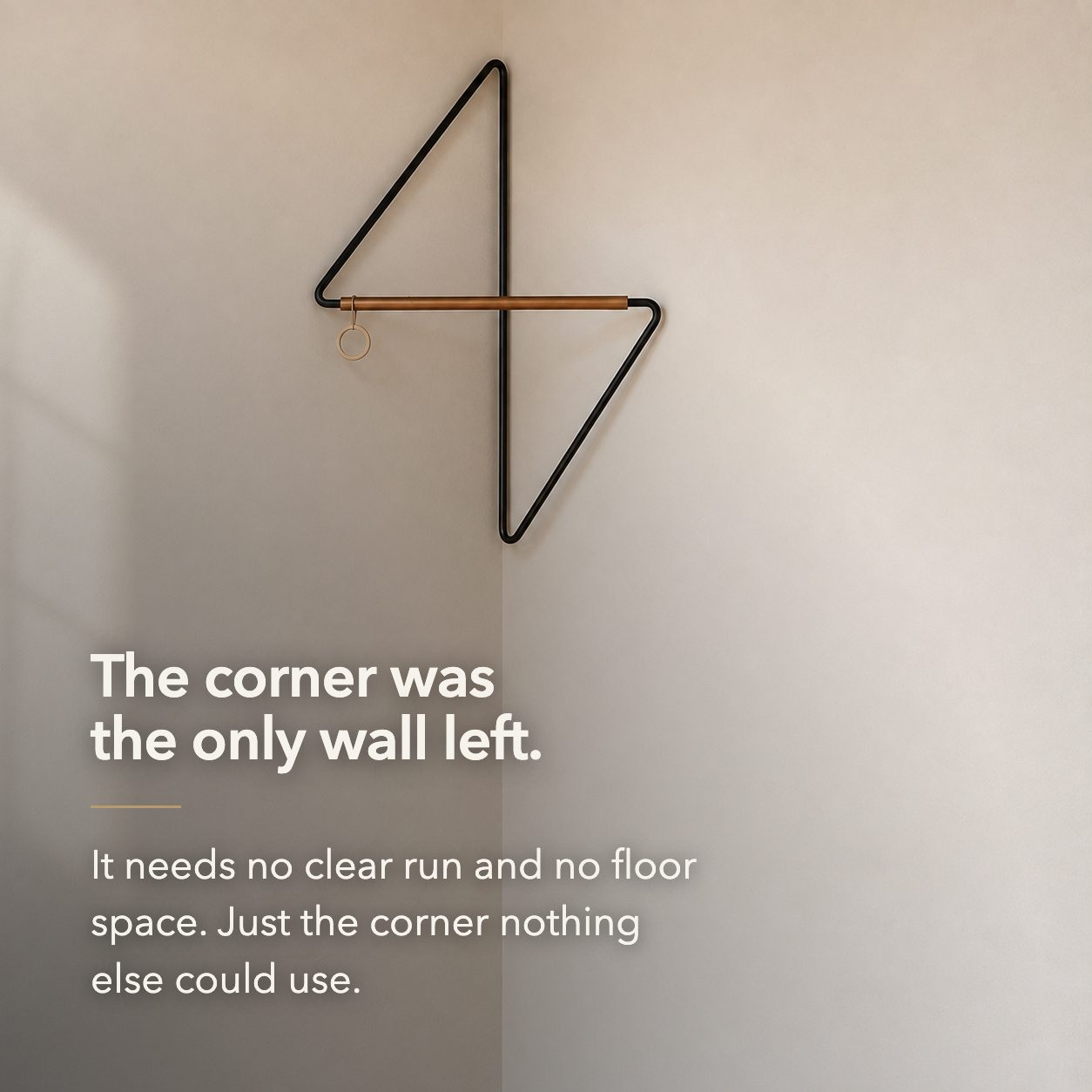 The bare black metal corner coat rack in a room corner, with overlaid text reading: The corner was the only wall left. It needs no clear run and no floor space.