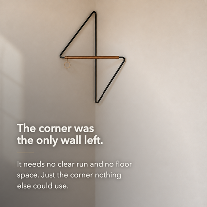 The bare black metal corner coat rack in a room corner, with overlaid text reading: The corner was the only wall left. It needs no clear run and no floor space.