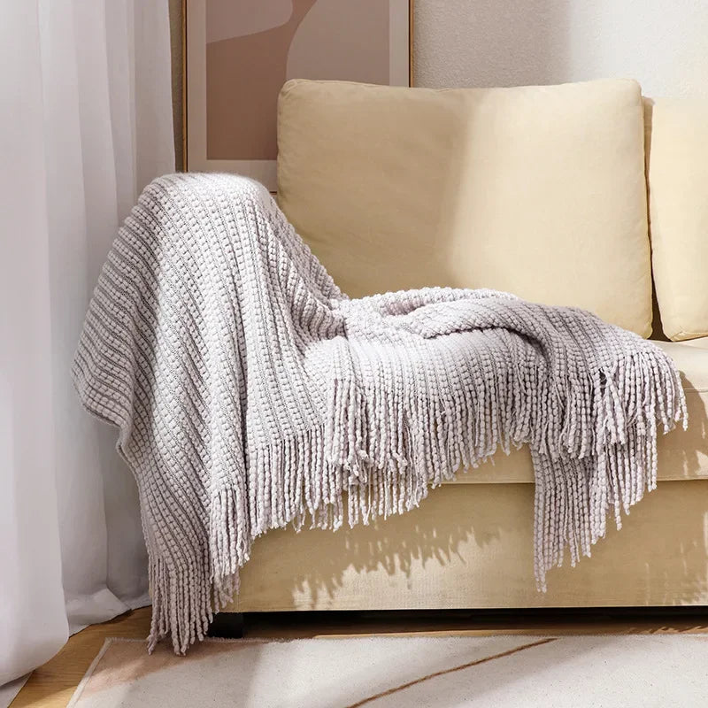 A gray bobble knit throw blanket with fringed ends draped over the arm of a sofa.