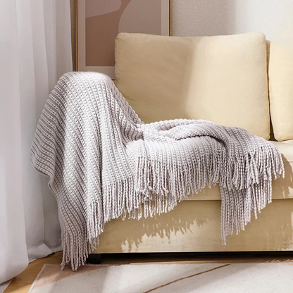 A gray bobble knit throw blanket with fringed ends draped over the arm of a sofa.
