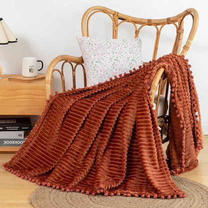 A rust flannel throw with raised stripes and matching pom-poms over a rattan armchair with a floral cushion