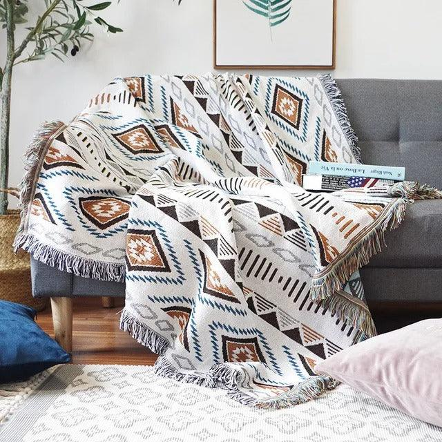 A white jacquard throw with brown and blue diamonds, thrown over a gray couch with fringe at the ends
