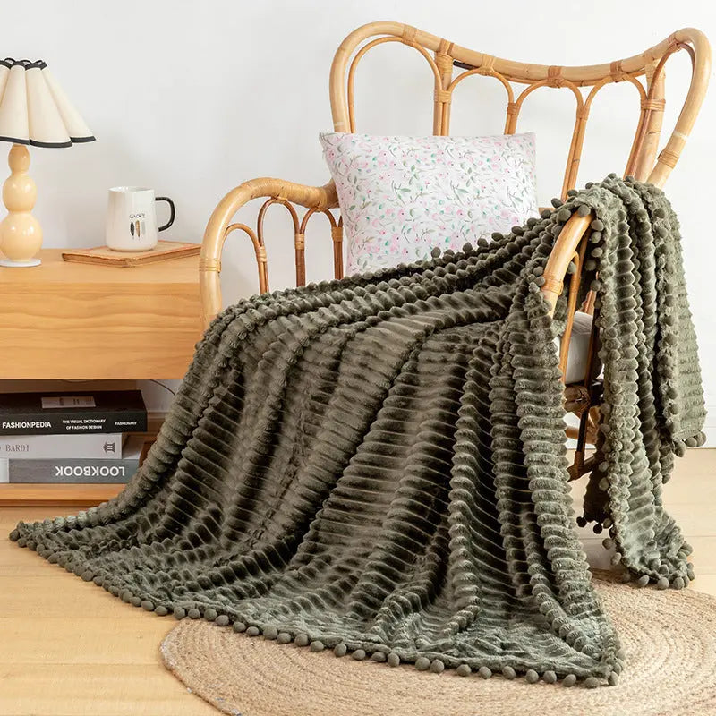 An olive flannel throw with raised stripes and an olive pom-pom edge over a rattan armchair