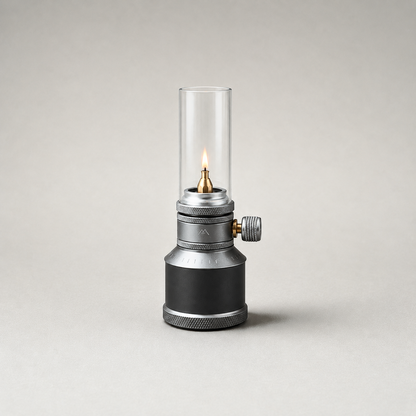 Front studio view of the lantern on a plain grey background, lit, showing the clear glass globe, the knurled collar with its flame knob, the gunmetal tank drum and the knurled base ring