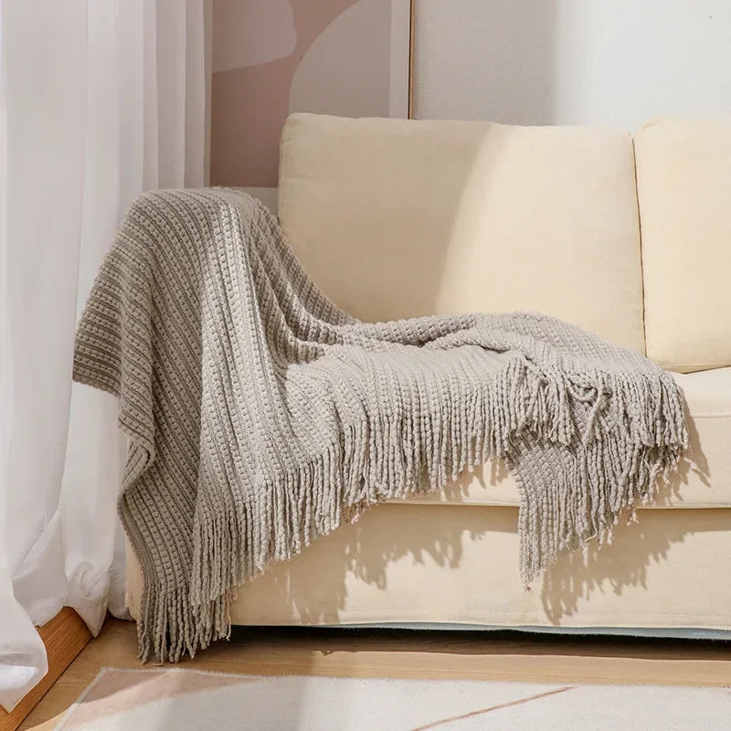 A taupe bobble knit throw blanket with fringed ends draped over the arm of a sofa.