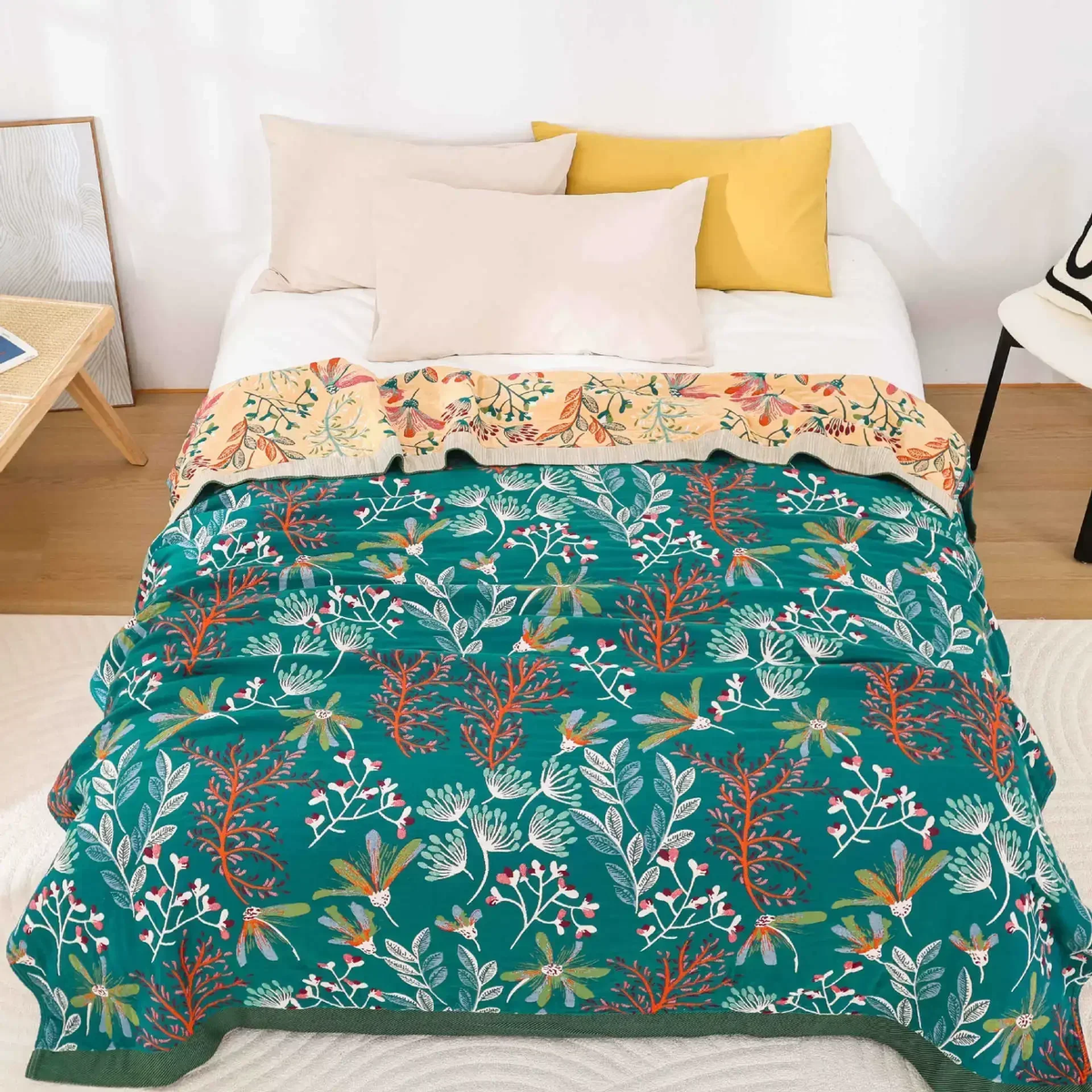 Teal wildflower gauze blanket spread over a bed, with the apricot floral reverse showing at the pillows and a dark green band along the foot