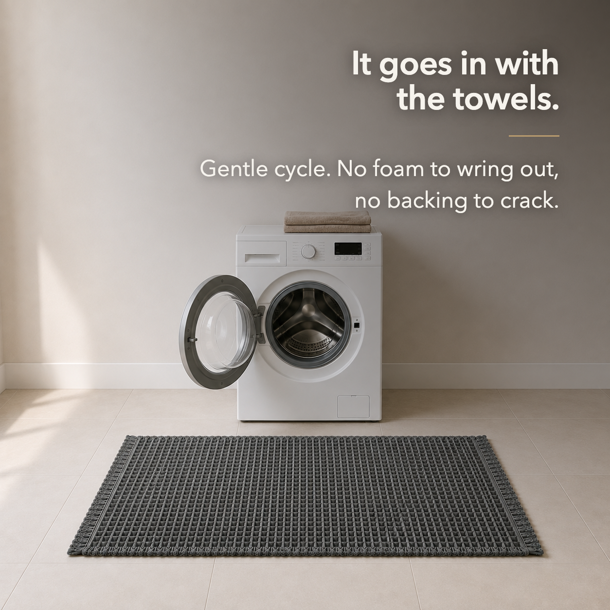 Charcoal waffle weave bath mat on a tiled floor in front of an open washing machine, captioned It goes in with the towels