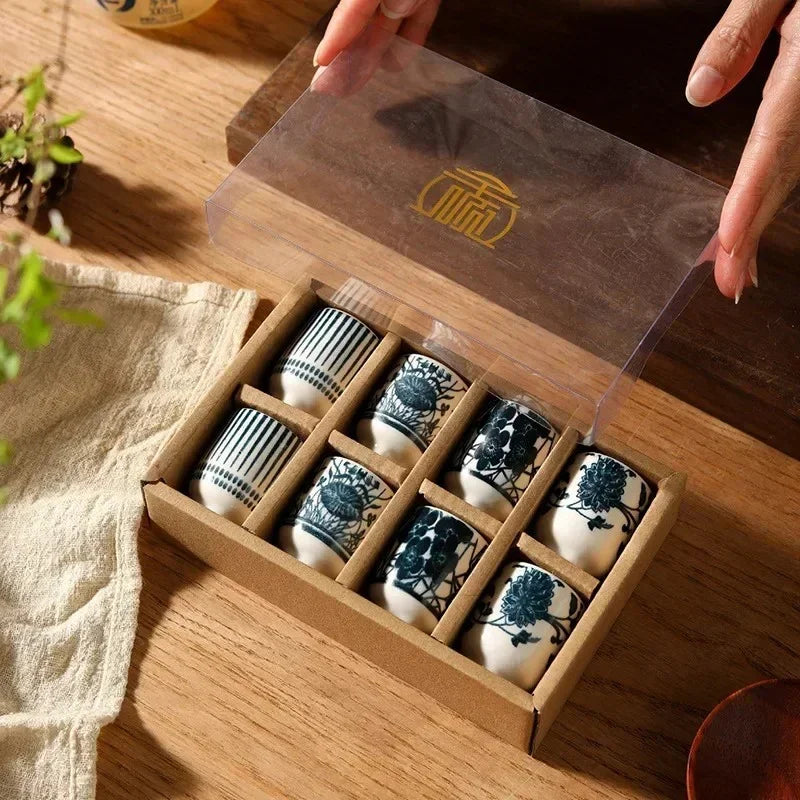 The eight-cup sake set in a kraft tray box, the clear lid lifted off, cups in pairs of four blue patterns.