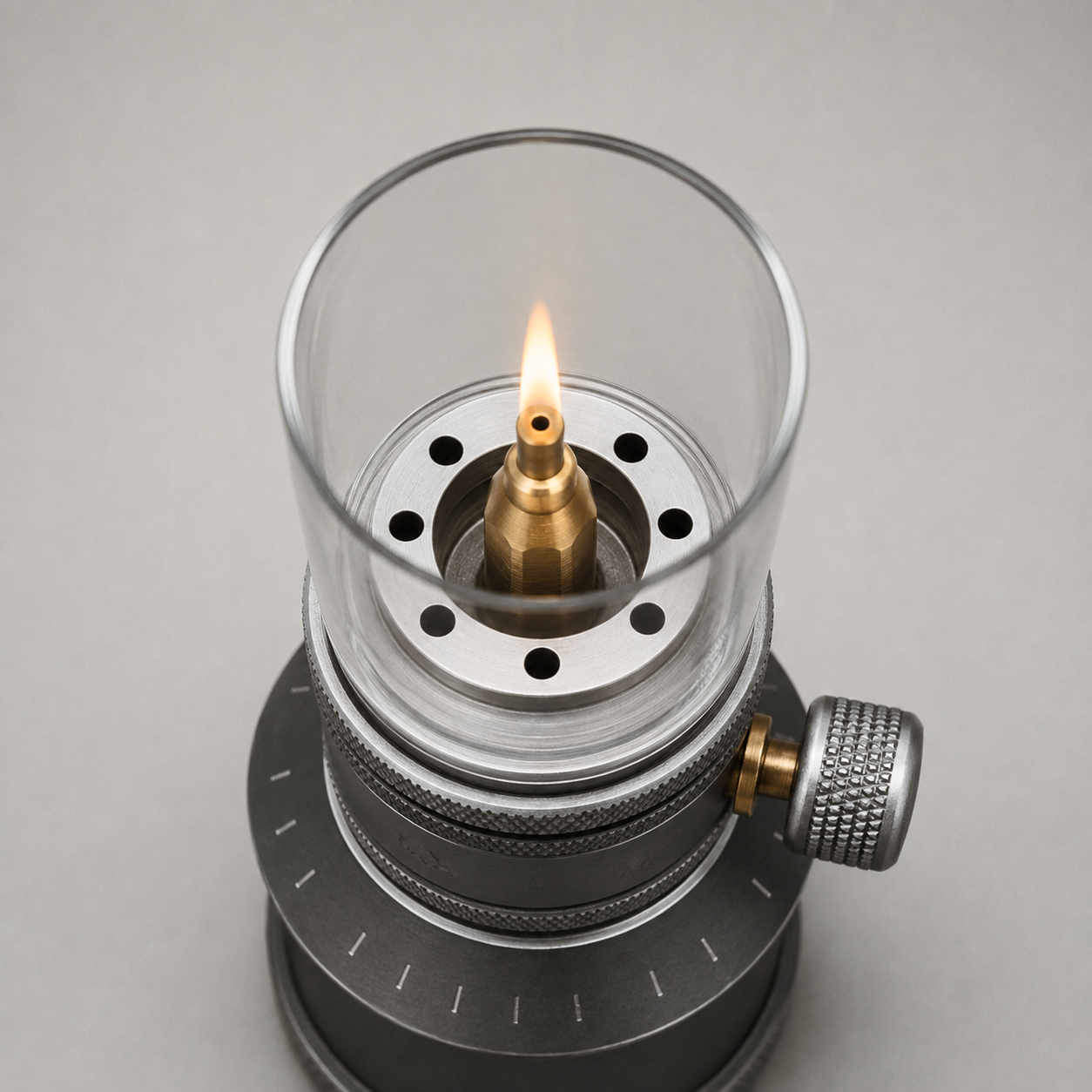 Close-up looking straight down into the open glass globe at the silver burner deck with its eight small round holes, and the stepped brass nozzle in the centre with the flame rising from the single hole in its top