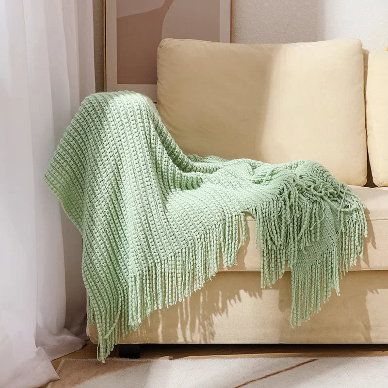 A sage bobble knit throw blanket with fringed ends draped over the arm of a sofa.