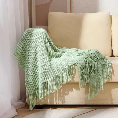 A sage bobble knit throw blanket with fringed ends draped over the arm of a sofa.