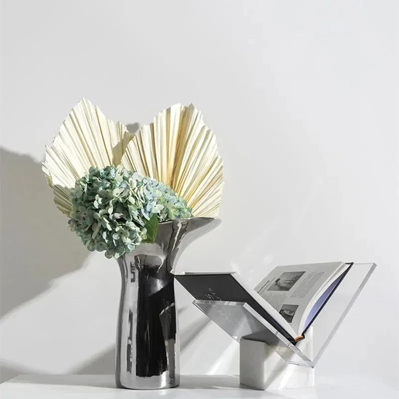 White marble stand holding an open book in its clear acrylic V, beside a chrome vase of dried leaves
