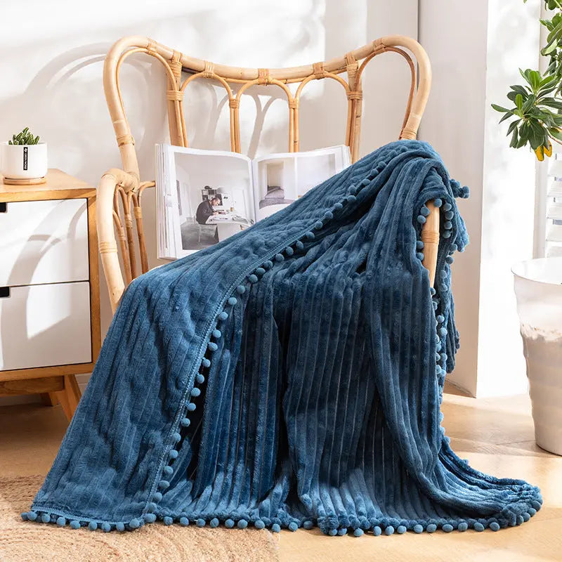 A blue flannel throw with raised stripes and blue pom-poms along the edge, over a rattan armchair