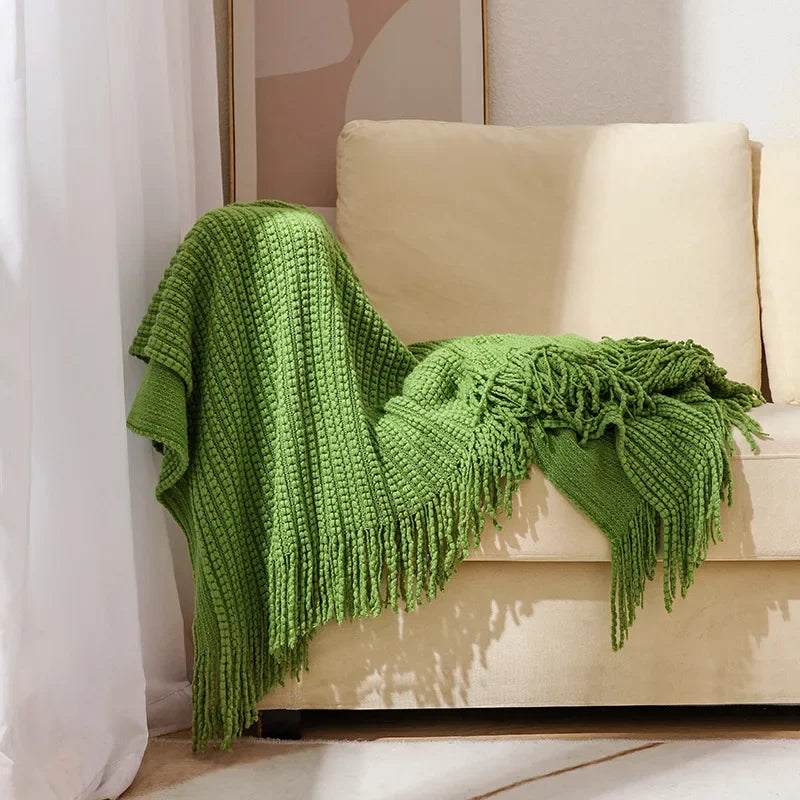A green bobble knit throw blanket with fringed ends draped over the arm of a sofa.
