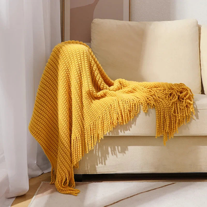 A mustard bobble knit throw blanket with fringed ends draped over the arm of a sofa.