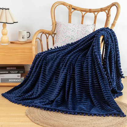 A navy flannel throw with raised stripes and navy pom-poms falling from a rattan armchair onto a jute rug