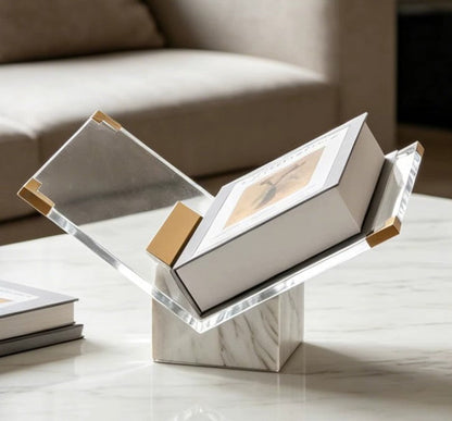 White and gold marble book stand with gold-toned corner caps holding a boxed book on a marble coffee table