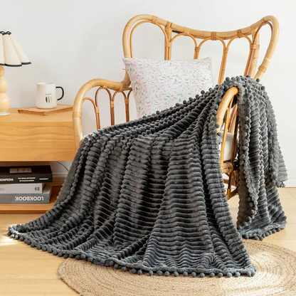 A charcoal flannel throw with raised stripes and charcoal pom-poms over a rattan armchair on a jute rug