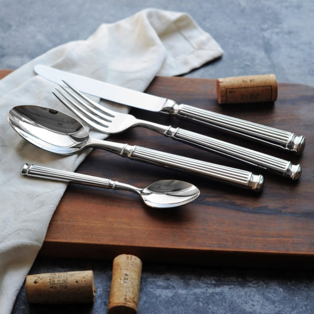 Five pieces of fluted stainless steel cutlery on a dark walnut board with wine corks and a white linen napkin