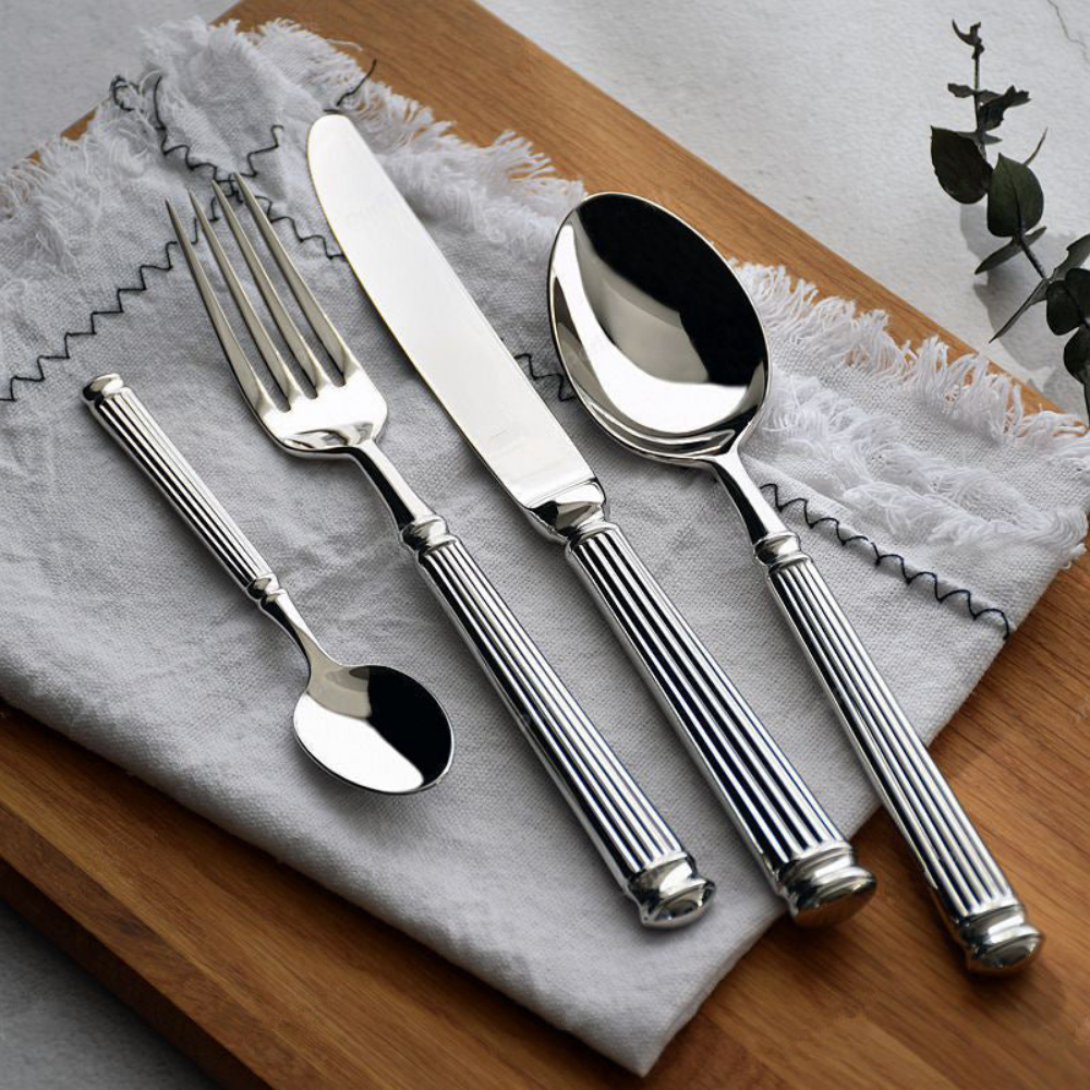 Five pieces of fluted stainless steel cutlery fanned across grey linen on a wooden board, the handles ending in stepped rounded caps